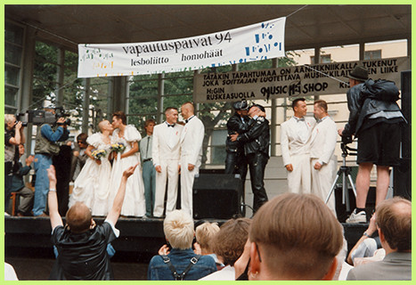 Four same-sex couples on stage. First on the left is a lesbian couple kissing, with matching white wedding dresses. The second couple is a gay couple with matching white suits. The third one is a gay couple in leather, kissing. The fourth one is a gay couple in matching suits, looking at each other. Above them is a banderoll with the name of the the event: Vapautuspäivät 1994, lesboliitto, homohäät - lesbian marriage, gay wedding.